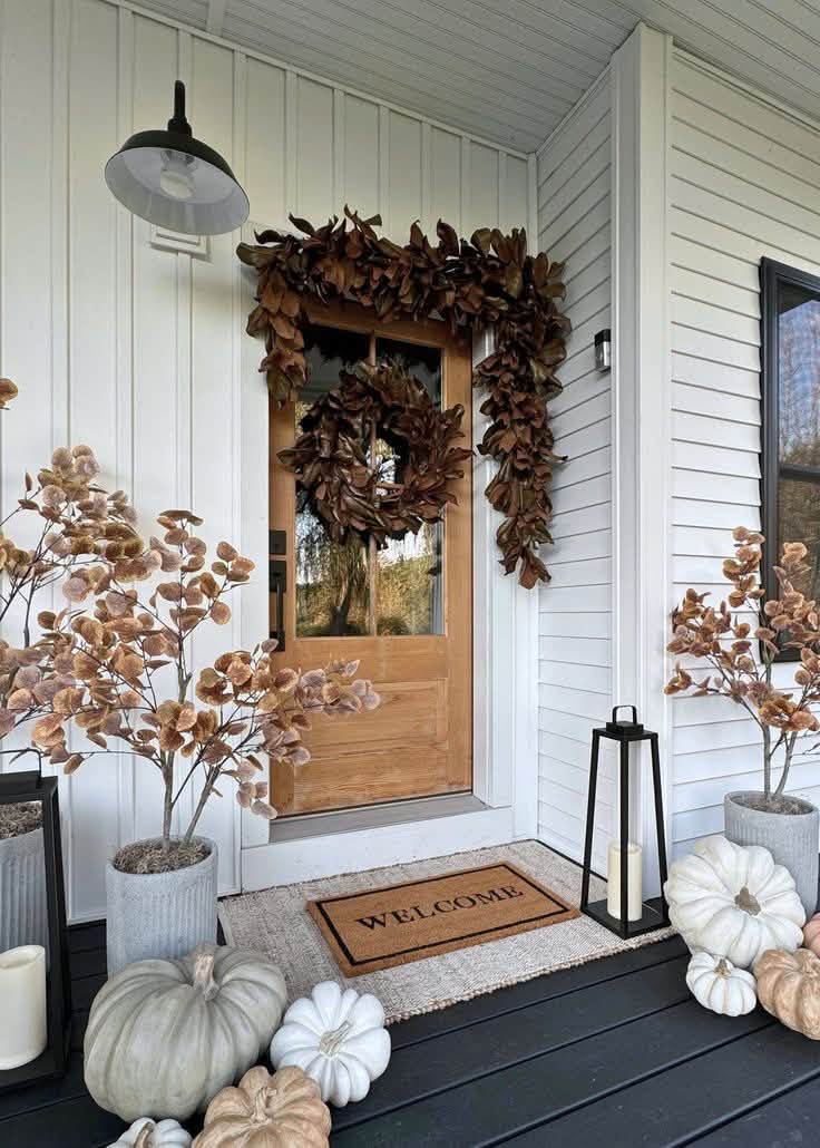 Wood front door framed with a magnolia garland, potted autumn stems, lanterns and white pumpkins
